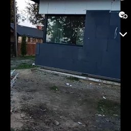 Jakub Skrzynski - Niedokończona elewacja budynku z widoczną warstwą ocieplenia w kolorze grafitowym i fragmentem białego styropianu, okno z odbiciem drzew, ziemia z resztkami materiałów budowlanych.