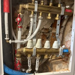 Jakub Skrzynski - Rozdzielacz ogrzewania podłogowego w szafce, widoczne zawory, rury doprowadzające i odprowadzające wodę z napisem 'ZASIL GRZEJNIKL' oraz 'PODŁOGA', zamontowany na tle surowej, częściowo obitej...