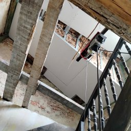 Jakub Skrzynski - Widok z dołu na strop w trakcie remontu, z widocznymi drewnianymi belkami, nową płytą gipsową podwieszoną na metalowych profilach, ociepleniem z wełny mineralnej oraz elektrycznym podnośnikiem...