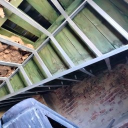Jakub Skrzynski - Konstrukcja sufitu podwieszanego z profili metalowych, ocieplona wełną mineralną, widoczna na tle starej ceglanej ściany i zielonej drewnianej podłogi poddasza.