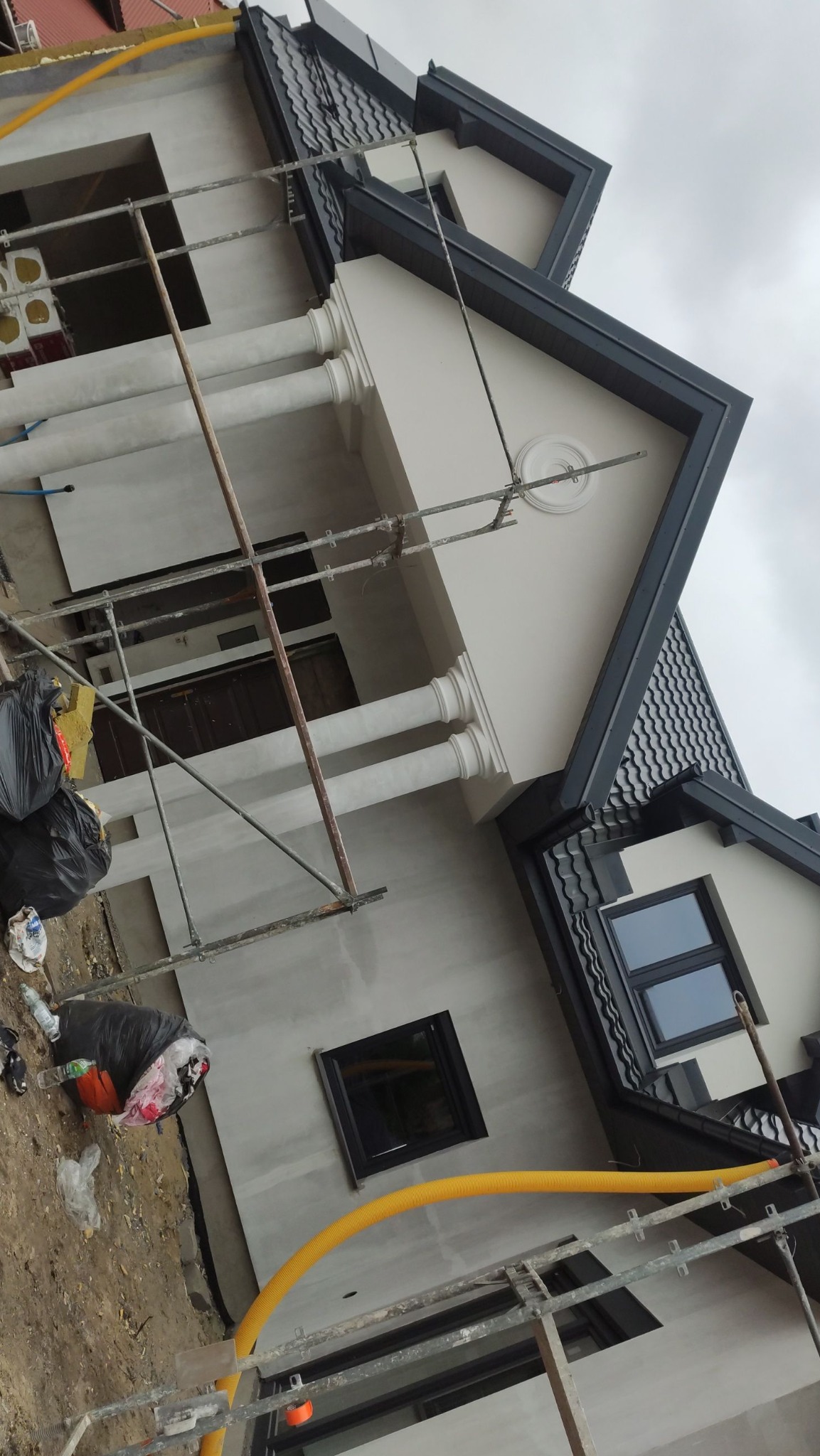 Niedokończona elewacja domu z widocznymi rusztowaniami, workami na śmieci i żółtym wężem wentylacyjnym, w tle pochmurne niebo.