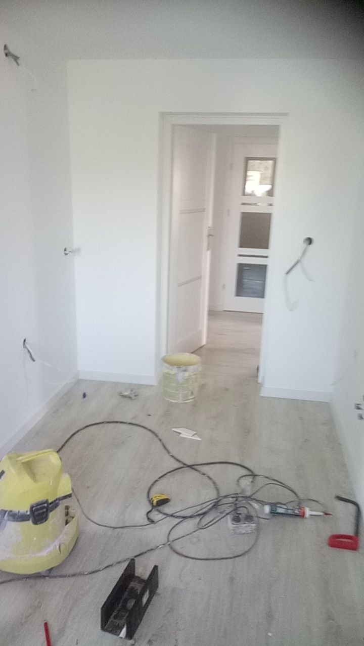Pokój w trakcie prac wykończeniowych: świeżo pomalowane białe ściany, widoczne przewody elektryczne, narzędzia i sprzęt budowlany na podłodze imitującej drewno, otwarte drzwi do kolejnego...