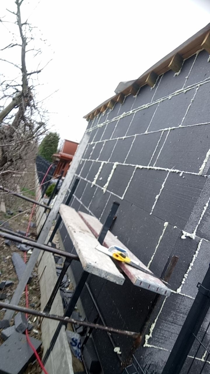 Ściana budynku pokryta czarnym styropianem, wypełniona pianką montażową, widoczne rusztowanie z deskami i narzędzia do cięcia.