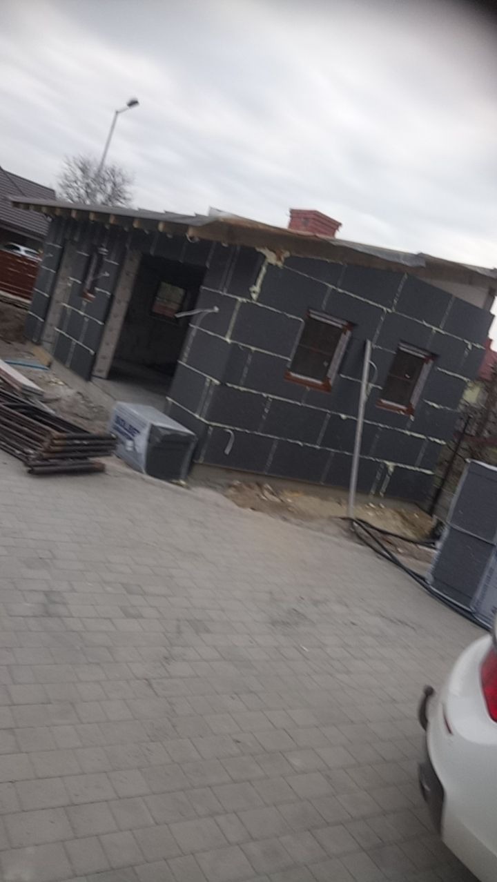 Budynek w trakcie ocieplania elewacji, widoczne płyty izolacyjne z szarym styropianem, okna z brązowymi ramami, przygotowane materiały budowlane oraz kostka brukowa na podjeździe.