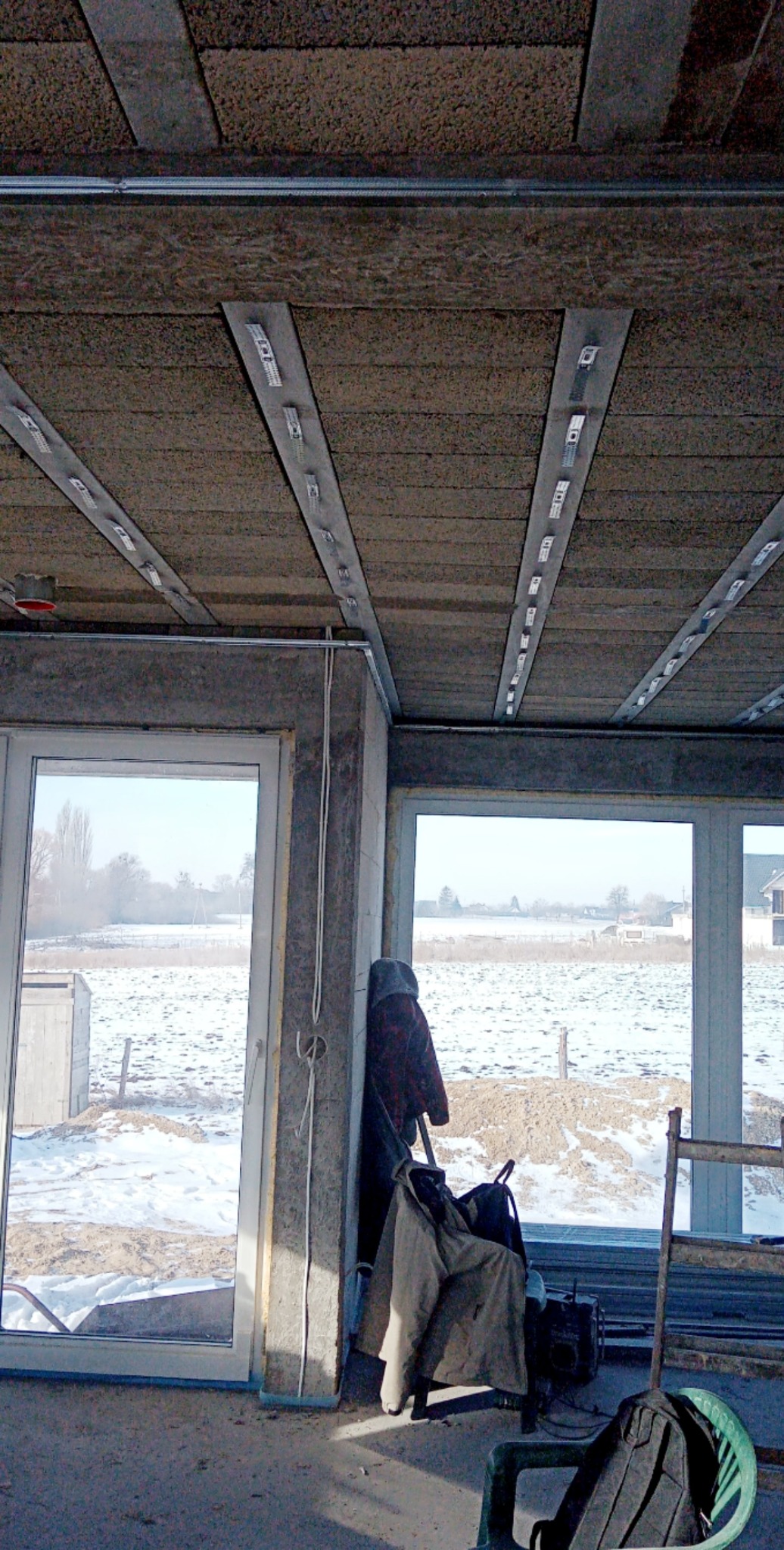 Montaż sufitu z paneli izolacyjnych w nowym budynku, widoczne metalowe profile i mocowania, okna z widokiem na zimowy krajobraz.