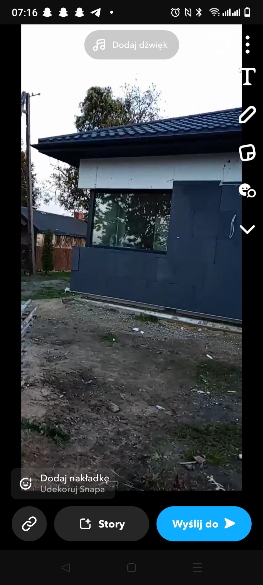 Niedokończona elewacja budynku z widoczną warstwą ocieplenia w kolorze grafitowym i fragmentem białego styropianu, okno z odbiciem drzew, ziemia z resztkami materiałów budowlanych.