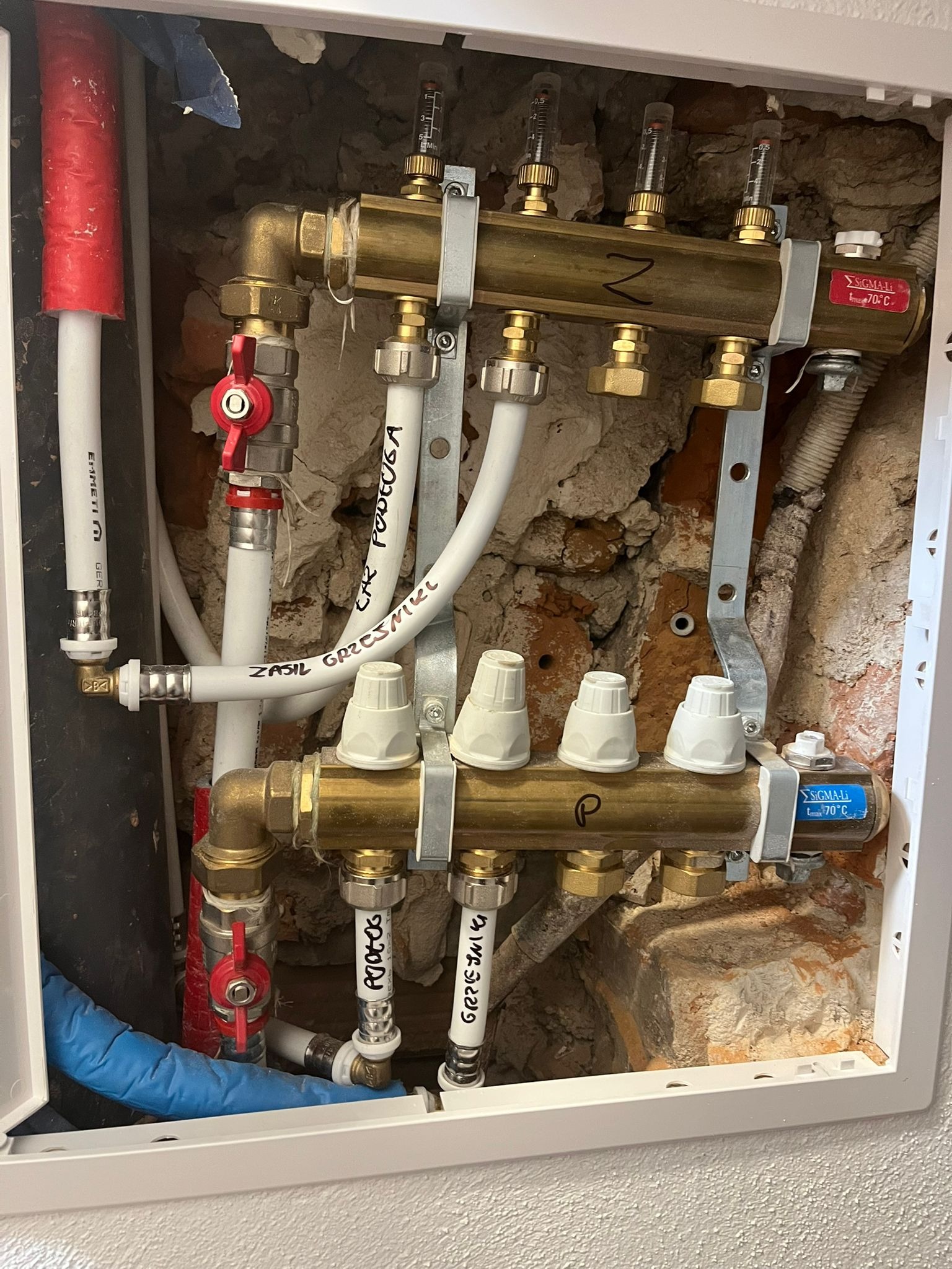 Rozdzielacz ogrzewania podłogowego w szafce, widoczne zawory, rury doprowadzające i odprowadzające wodę z napisem 'ZASIL GRZEJNIKL' oraz 'PODŁOGA', zamontowany na tle surowej, częściowo obitej...