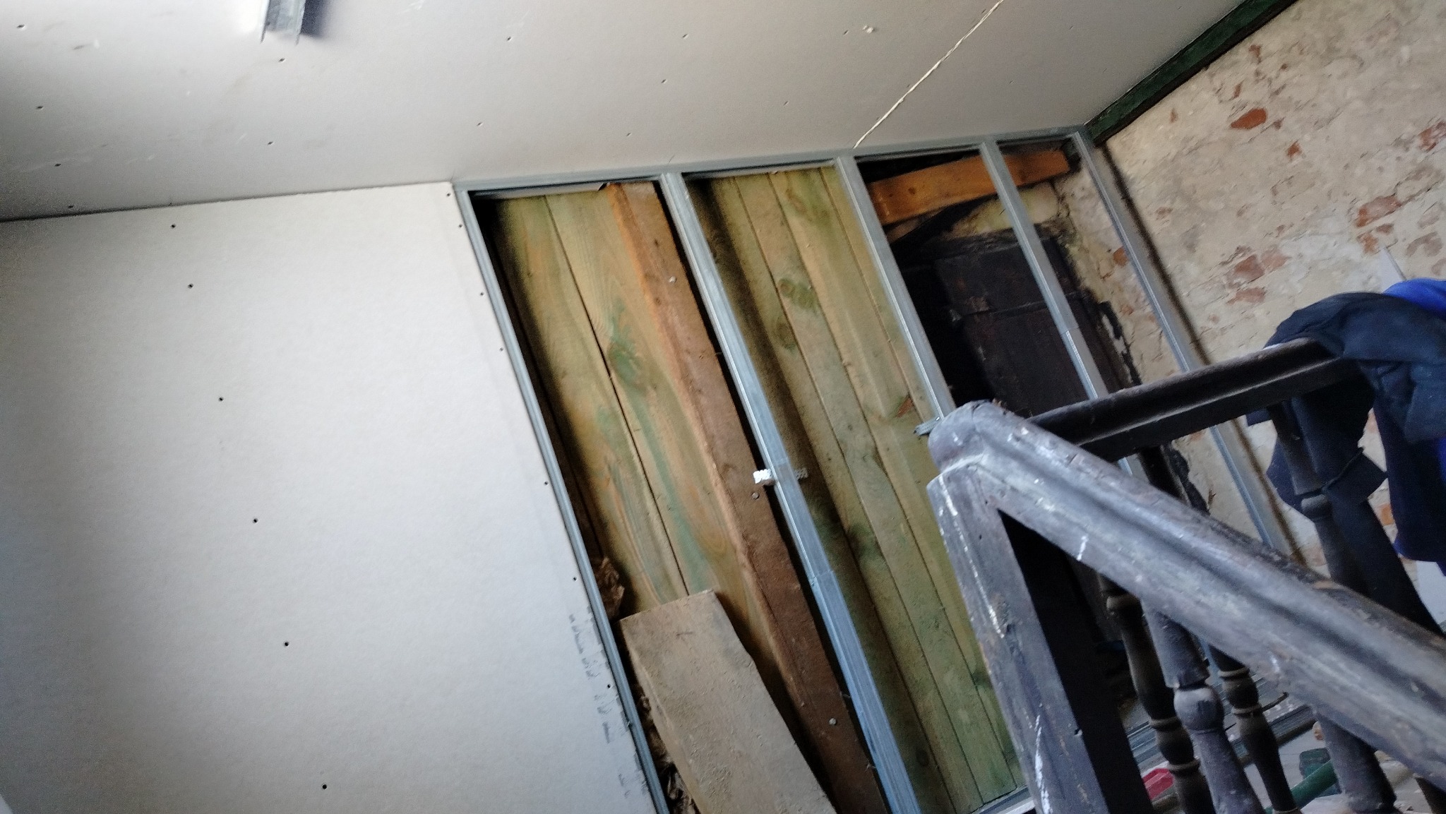Wnętrze w trakcie prac: płyta gipsowa na ścianie, metalowy stelaż, fragment starej ściany z cegły i drewniane elementy konstrukcyjne, widoczne schody z balustradą.