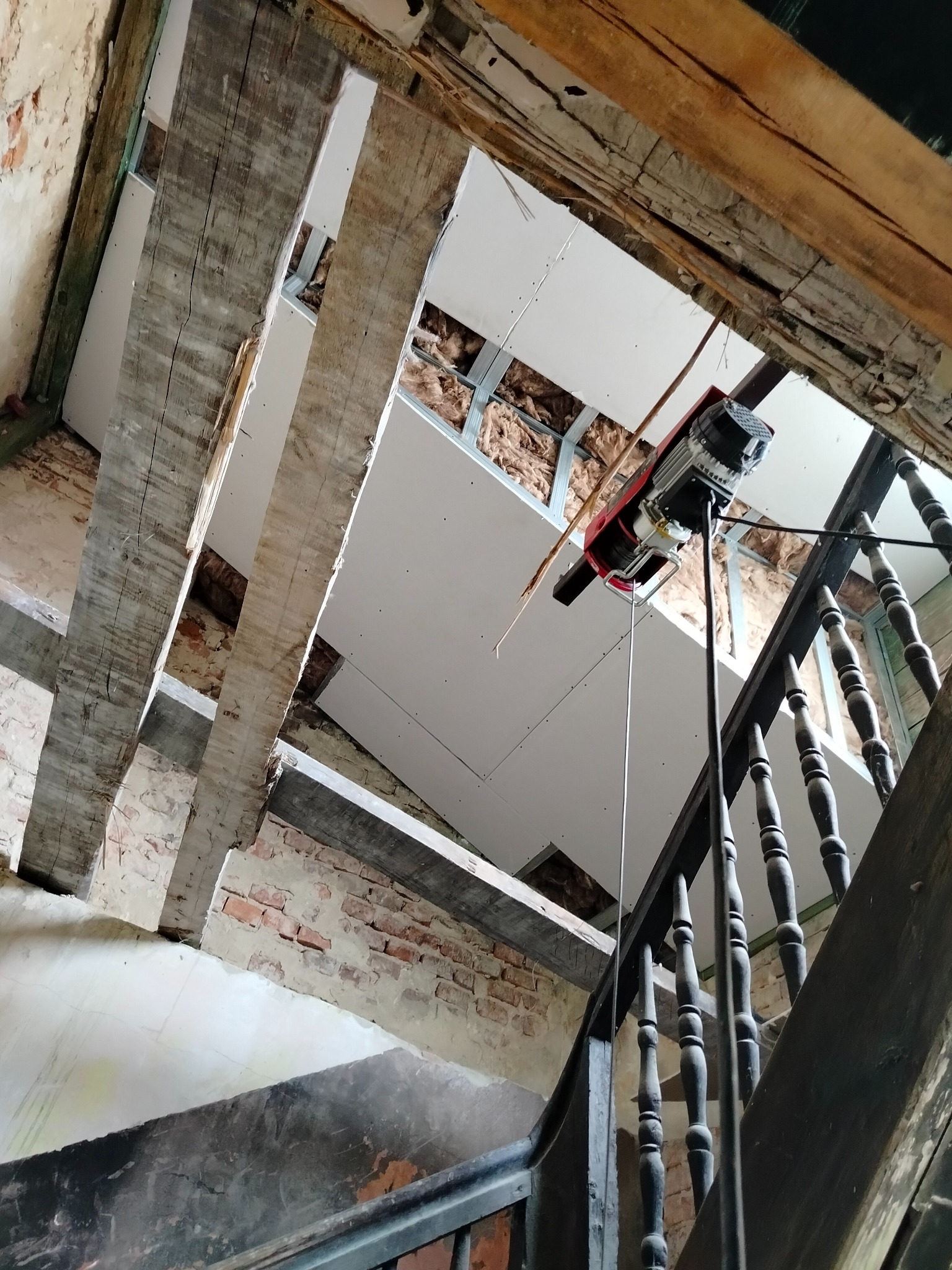 Widok z dołu na strop w trakcie remontu, z widocznymi drewnianymi belkami, nową płytą gipsową podwieszoną na metalowych profilach, ociepleniem z wełny mineralnej oraz elektrycznym podnośnikiem...