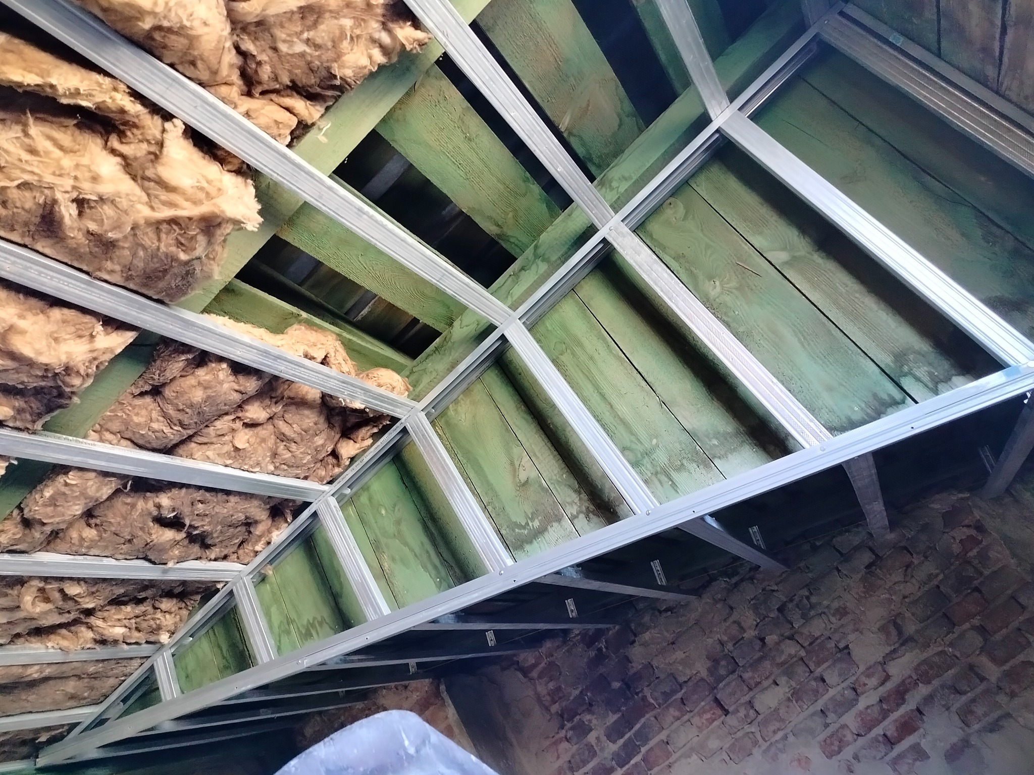 Izolacja poddasza wełną mineralną z konstrukcją metalową, widoczne zielone deski i fragment murowanej ściany.