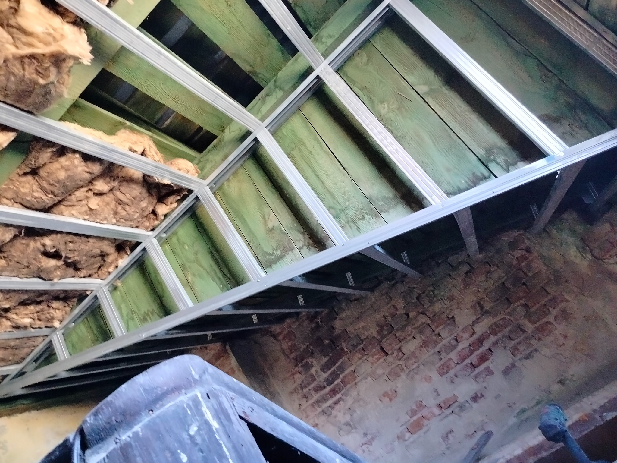 Konstrukcja sufitu podwieszanego z profili metalowych, ocieplona wełną mineralną, widoczna na tle starej ceglanej ściany i zielonej drewnianej podłogi poddasza.