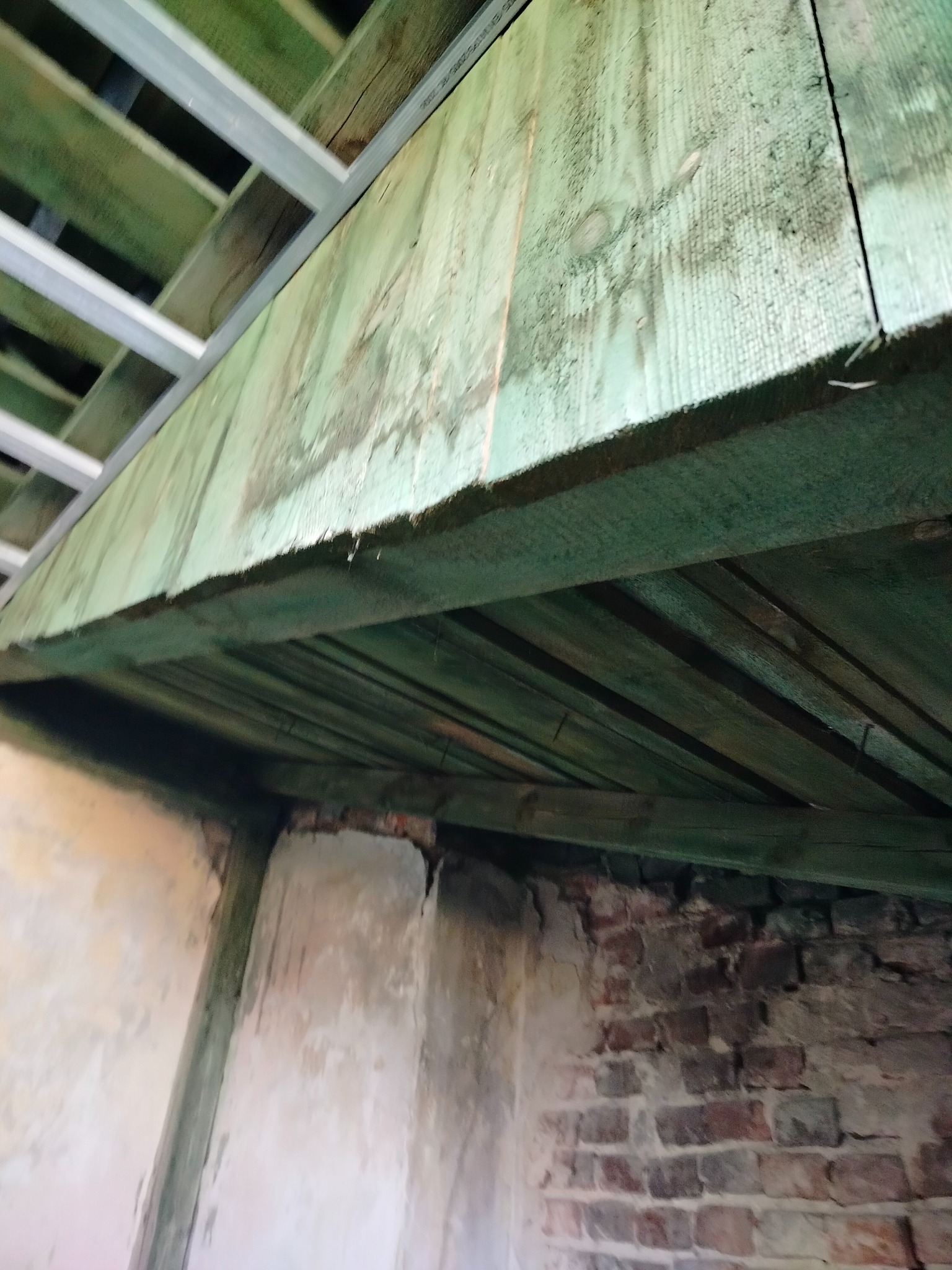 Surowy stan poddasza z widoczną drewnianą konstrukcją stropu pomalowaną na zielono, metalowymi profilami sufitowymi i fragmentem ściany z cegły.