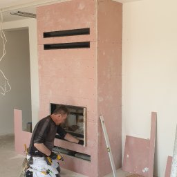 DaJF MAKSYMILIAN BOREK - Pracownik w trakcie obudowy kominka płytami kartonowo-gipsowymi, widoczne narzędzia, poziomica i fragmenty płyt na podłodze.