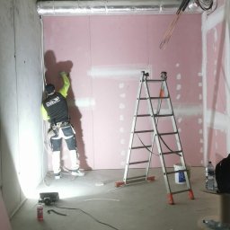 DaJF MAKSYMILIAN BOREK - Pracownik w odzieży roboczej szpachluje łączenia płyt karton-gips na ścianie w surowym pomieszczeniu z widoczną instalacją wentylacyjną pod sufitem, drabina aluminiowa obok.