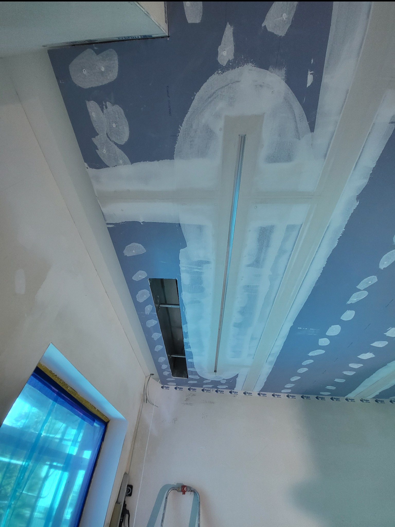 Montaż sufitu podwieszanego z płyt kartonowo-gipsowych, widoczne profile i wstępnie zaszpachlowane łączenia, fragment okna zabezpieczonego folią.