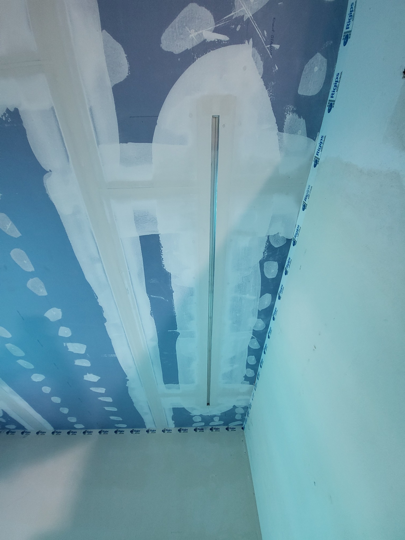 Montaż sufitu podwieszanego: widok na częściowo zaszpachlowaną płytę kartonowo-gipsową z widocznymi łączeniami wzmocnionymi taśmą, metalowy profil sufitowy zamontowany w centralnej części, płyta...