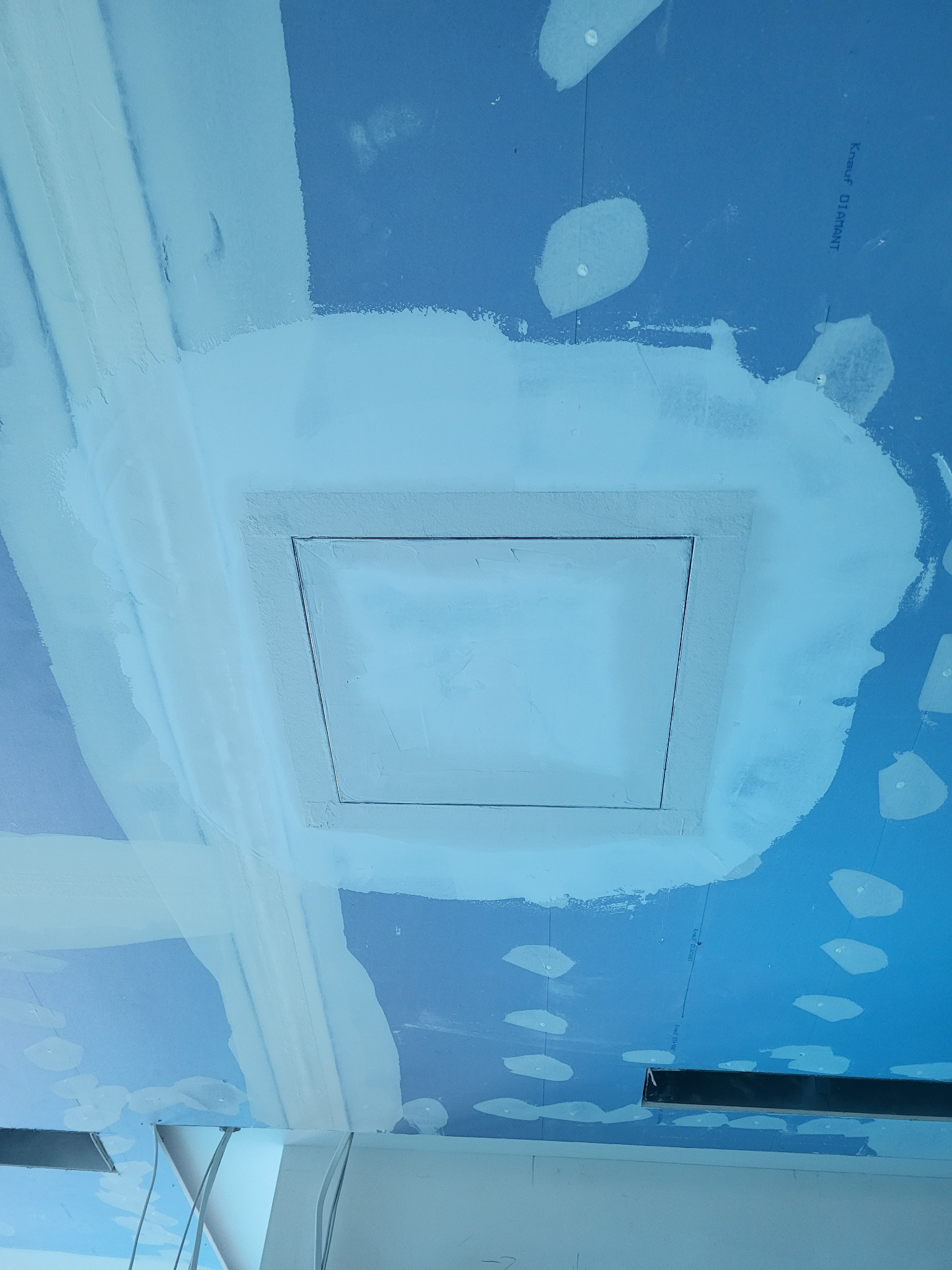 Niebieska płyta karton-gips z zamontowaną klapą rewizyjną, otoczona świeżo nałożoną gładzią szpachlową, widoczne ślady po wkrętach i łączeniach płyt.