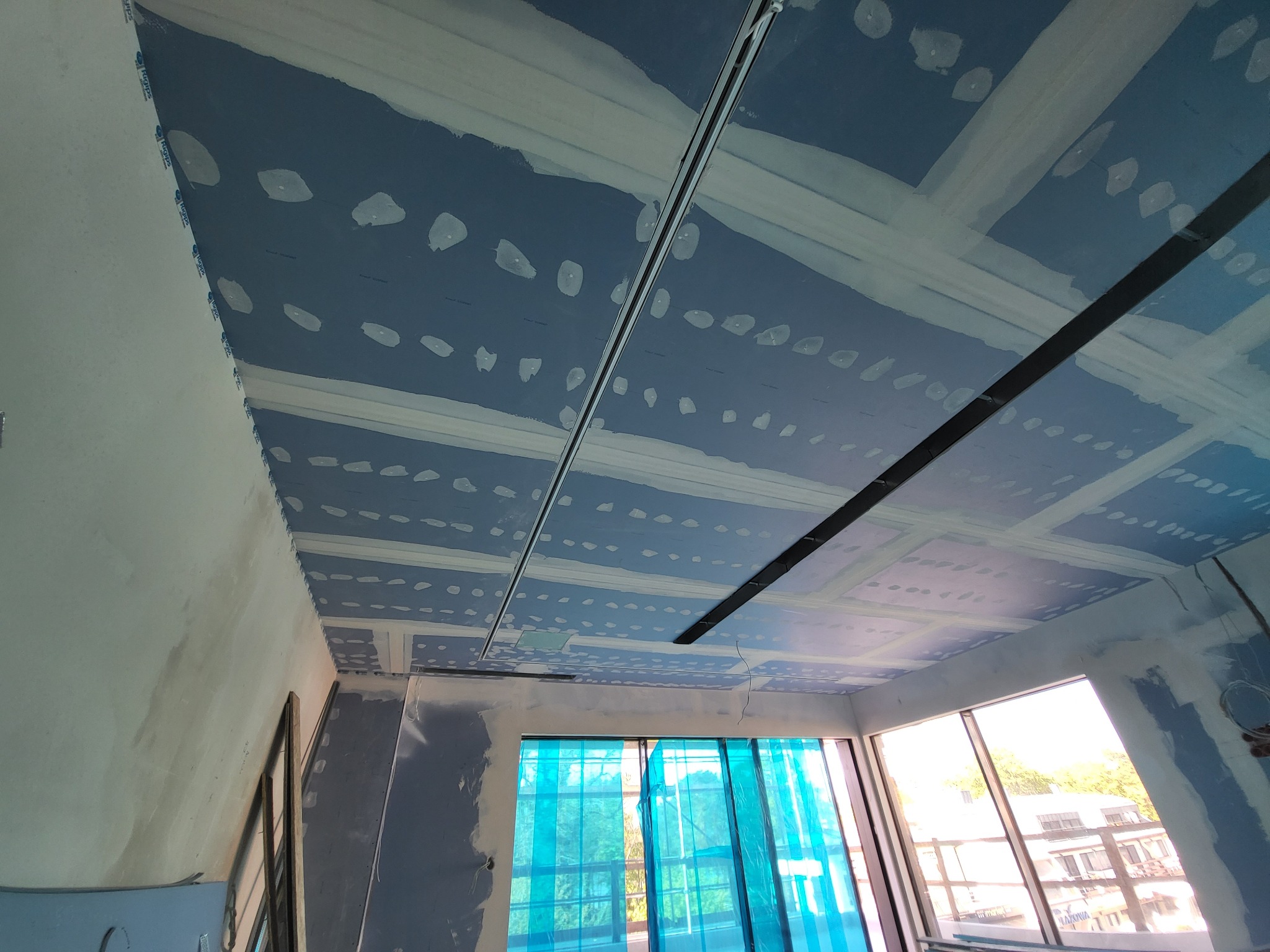 Niebieska płyta sufitowa z widocznymi łączeniami i elementami konstrukcyjnymi, częściowo pokryta białą masą szpachlową, okna zabezpieczone folią ochronną.