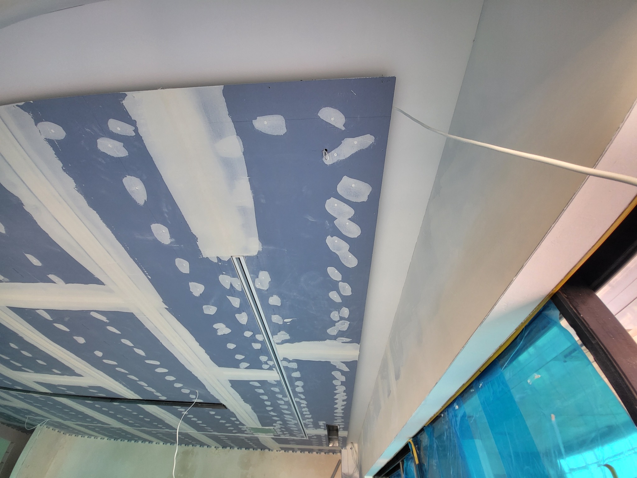 Sufit z zamontowaną konstrukcją pod płytę gipsowo-kartonową, widoczne punkty mocowania i łączenia płyt, częściowo pokryte masą szpachlową, zabezpieczone okno folią ochronną.