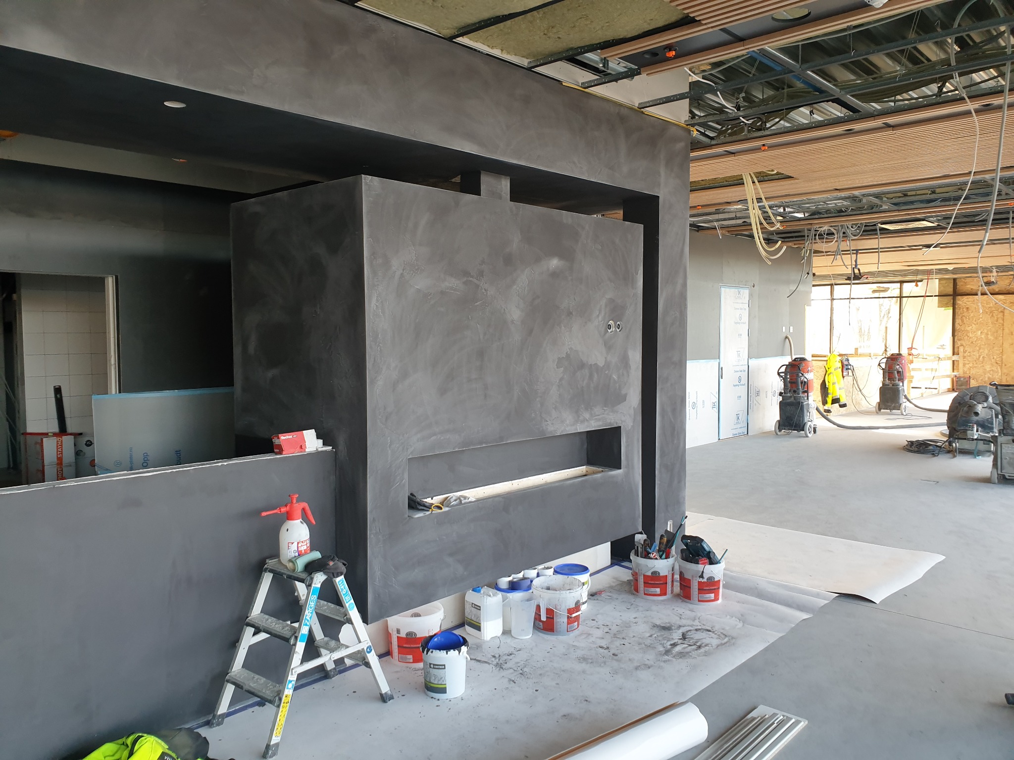 Wnętrze w trakcie prac wykończeniowych w Goleniowie: ściana z szarą, strukturalną powłoką, drabina z rozpylaczem, wiadra z farbą, widoczny sufit z instalacjami.