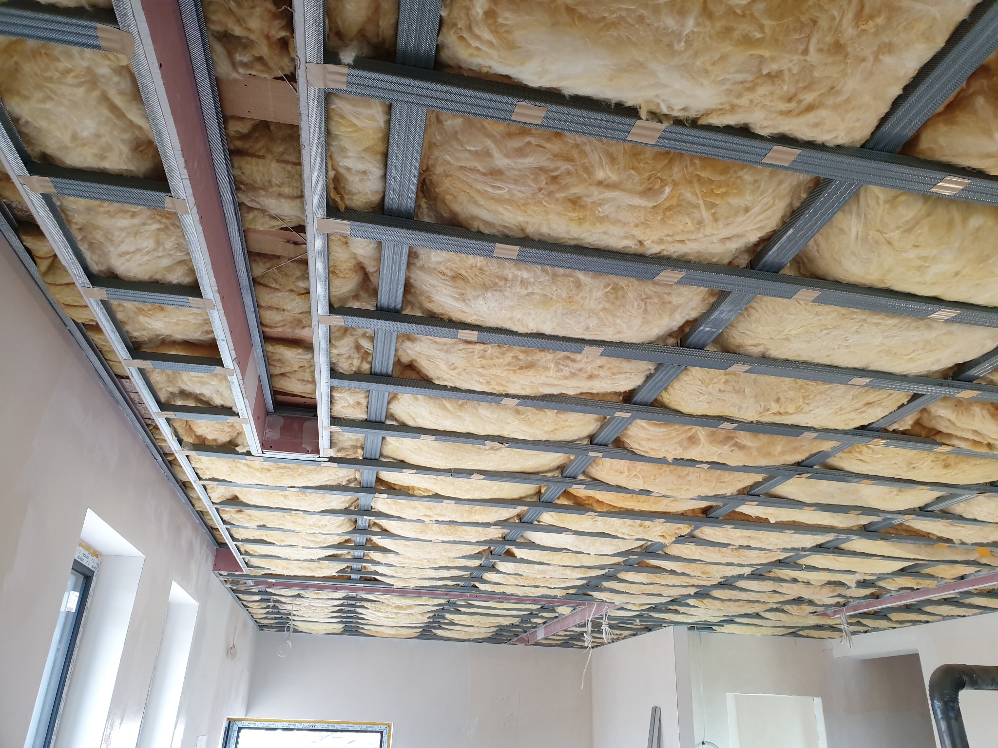Montaż izolacji z wełny mineralnej w metalowym stelażu podwieszanego sufitu w trakcie prac wykończeniowych.