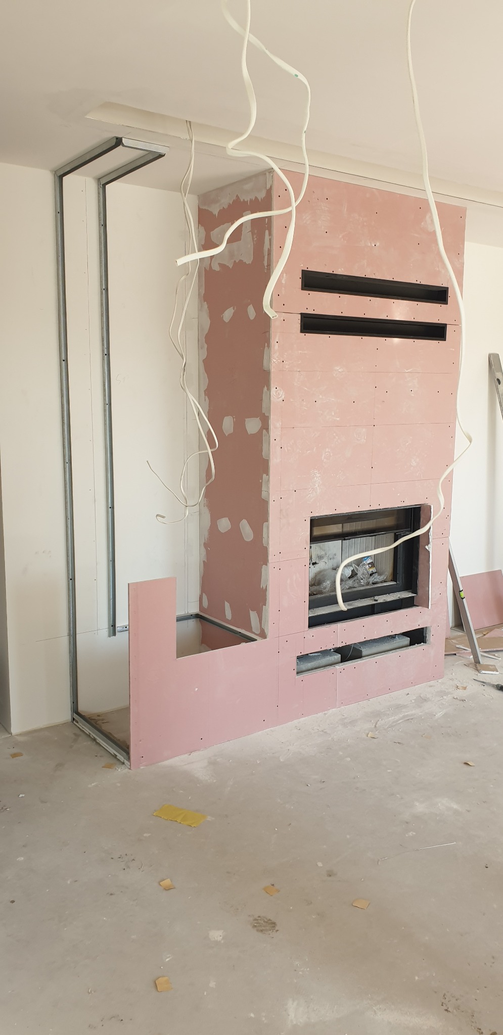Wnętrze w trakcie budowy kominka z obudową z płyt kartonowo-gipsowych, widoczne metalowe profile, instalacja elektryczna i otwory wentylacyjne w obudowie.