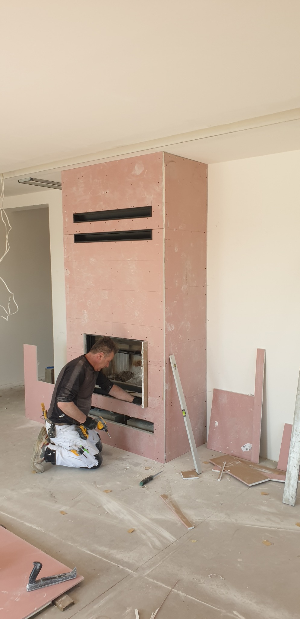 Pracownik w trakcie obudowy kominka płytami kartonowo-gipsowymi, widoczne narzędzia, poziomica i fragmenty płyt na podłodze.