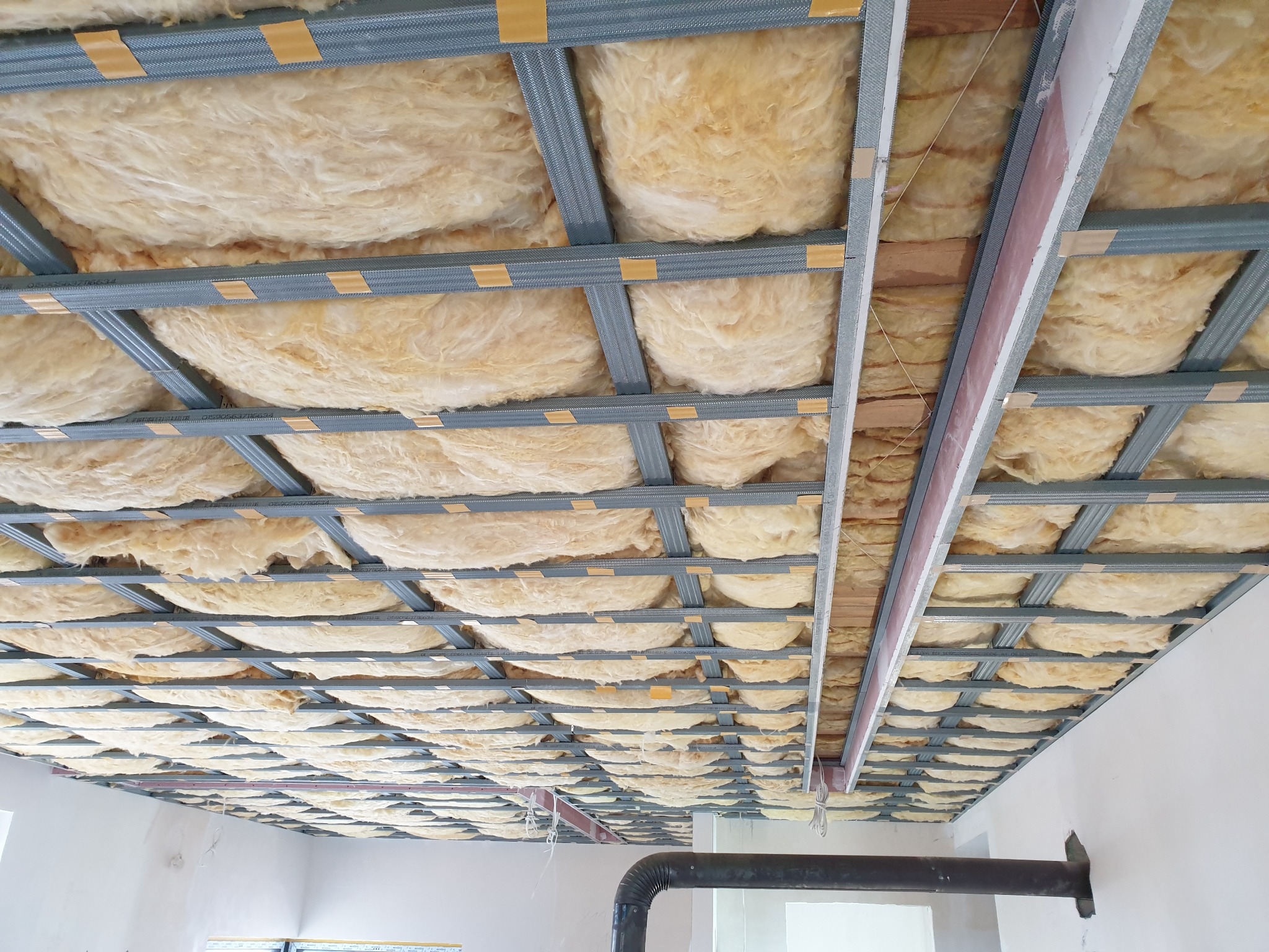 Metalowy stelaż sufitowy z wypełnieniem z wełny mineralnej, widok z dołu, fragment instalacji wentylacyjnej przy ścianie.