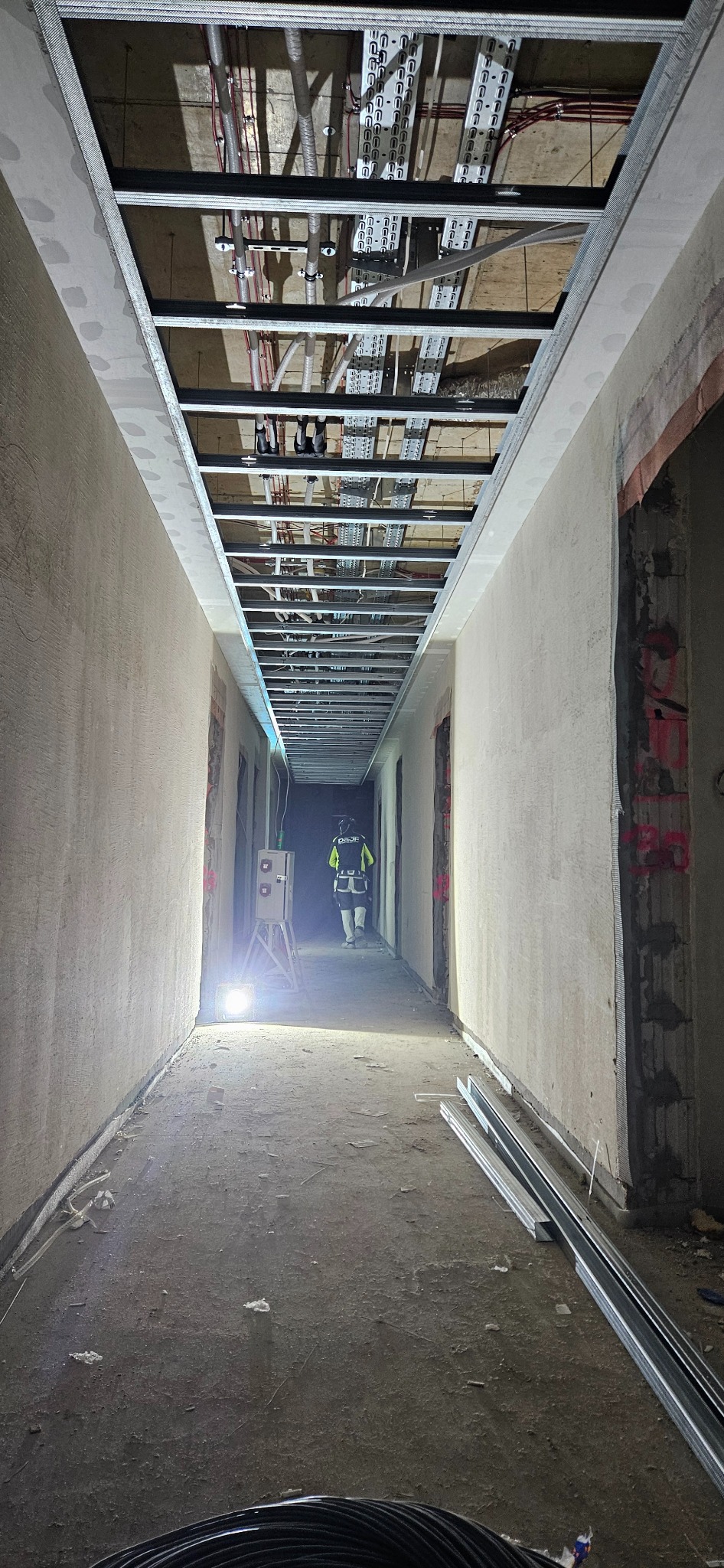Korytarz w trakcie prac remontowych z widoczną konstrukcją metalową sufitu podwieszanego, instalacjami i pracownikiem w oddali oświetlonym lampą.