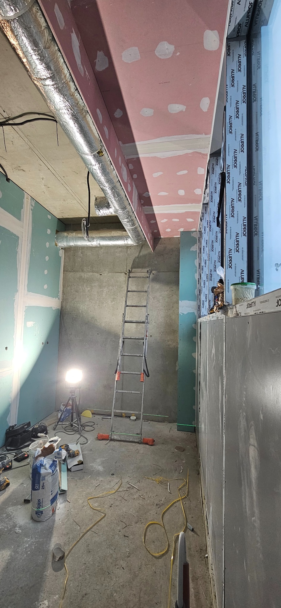 Wnętrze w trakcie prac wykończeniowych: widoczne płyty gipsowo-kartonowe na ścianach i suficie, aluminiowe rury wentylacyjne pod sufitem, drabina rozstawiona na środku pomieszczenia, narzędzia...
