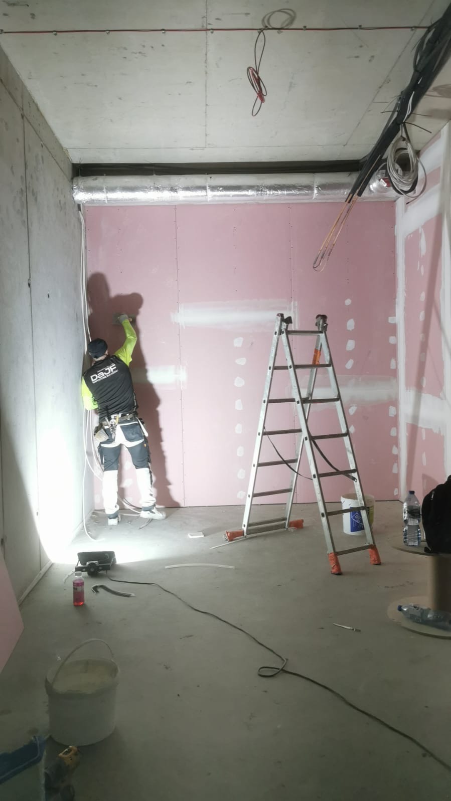 Pracownik w odzieży roboczej szpachluje łączenia płyt karton-gips na ścianie w surowym pomieszczeniu z widoczną instalacją wentylacyjną pod sufitem, drabina aluminiowa obok.