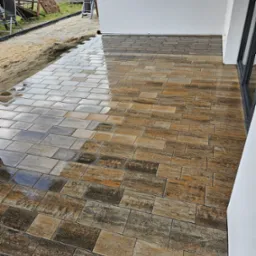 Ułożona kostka brukowa imitująca drewno, mokra od deszczu, na tarasie w Skierniewicach. W tle plac zabaw i przygotowany teren pod dalsze prace.