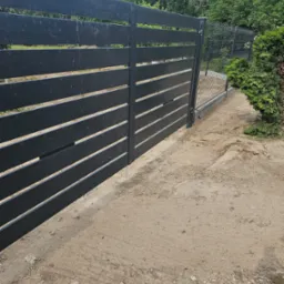 Nowoczesne, poziome ogrodzenie panelowe w kolorze grafitowym, z widoczną siatką ogrodzeniową w tle, na piaszczystym podłożu.