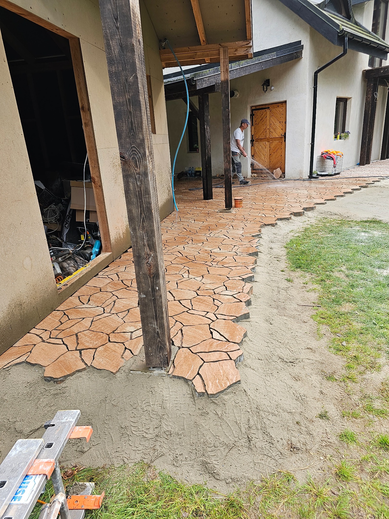 Układanie nieregularnej kostki brukowej w odcieniach brązu na podwórku, pracownik spłukuje powierzchnię wodą z węża, widoczny fragment domu z drewnianymi elementami i drabina oparta o ścianę.