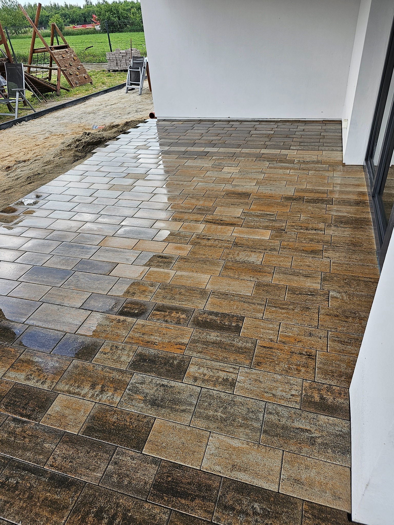 Ułożona kostka brukowa imitująca drewno, mokra od deszczu, na tarasie w Skierniewicach. W tle plac zabaw i przygotowany teren pod dalsze prace.