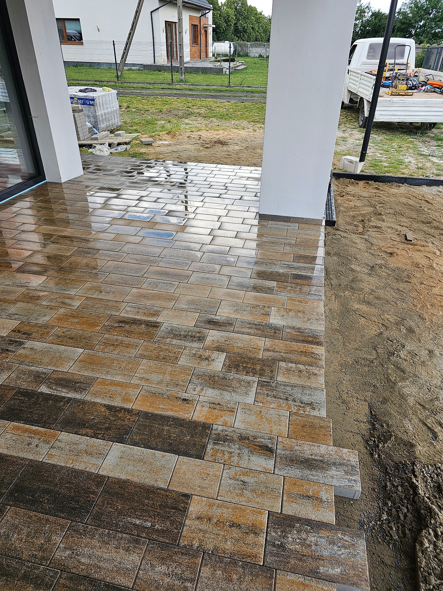 Świeżo ułożona, mokra kostka brukowa w odcieniach brązu i szarości, przylegająca do białego filaru werandy, obok przygotowany teren pod dalsze prace.