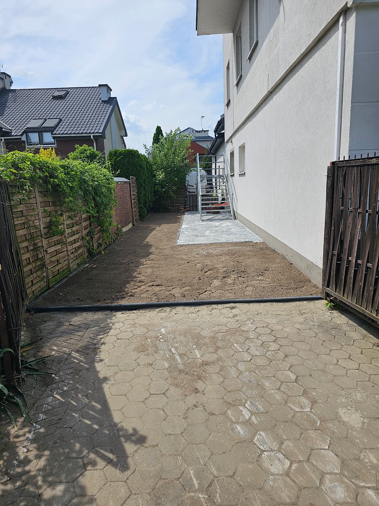 Przygotowany teren pod budowę ogrodzenia, widoczne obrzeża, częściowo ułożona kostka brukowa, budynek mieszkalny w tle.