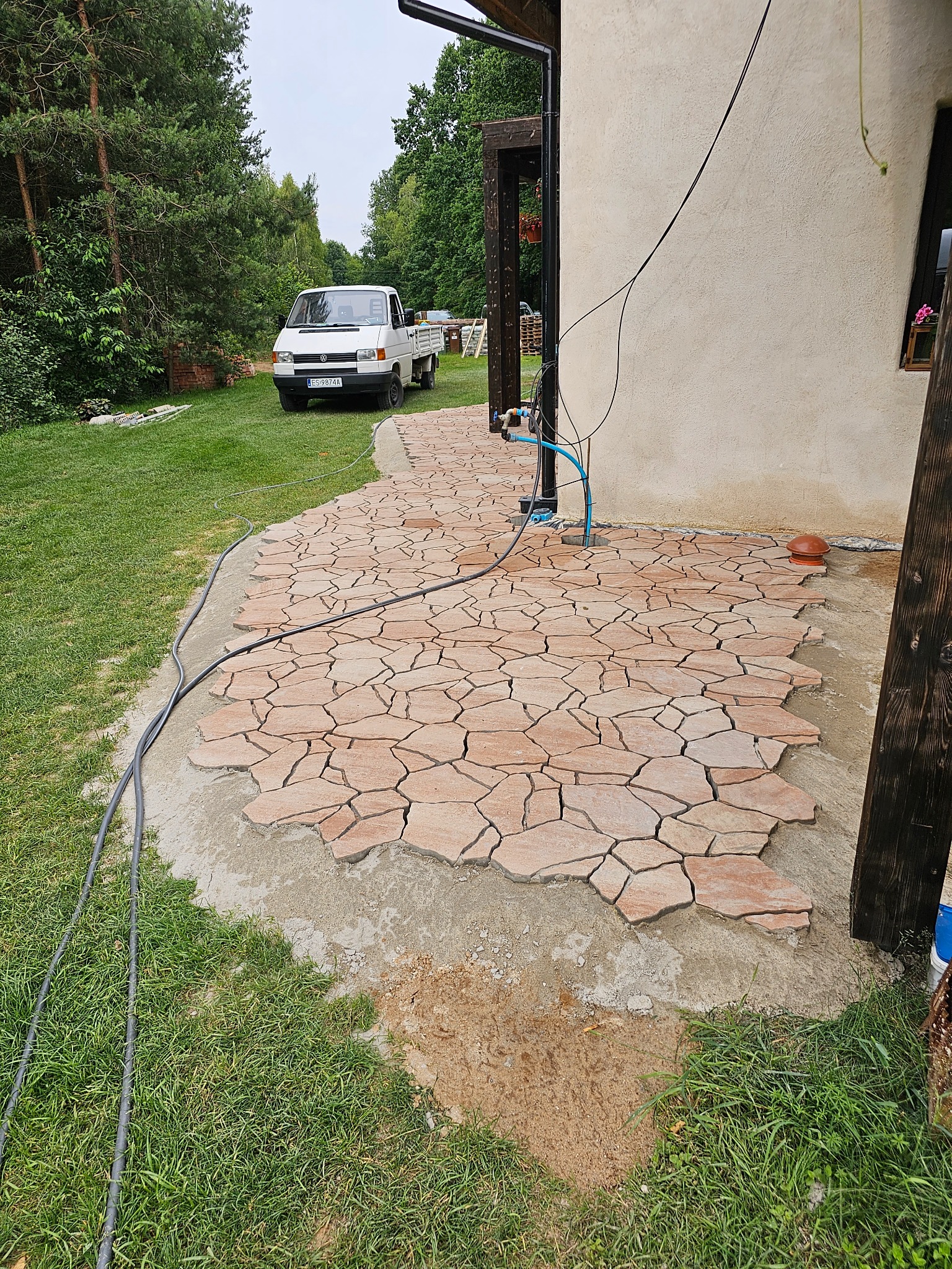 Nieregularny chodnik z czerwono-brązowych płyt kamiennych układany przy budynku, widoczny biały dostawczy samochód na trawniku w tle.
