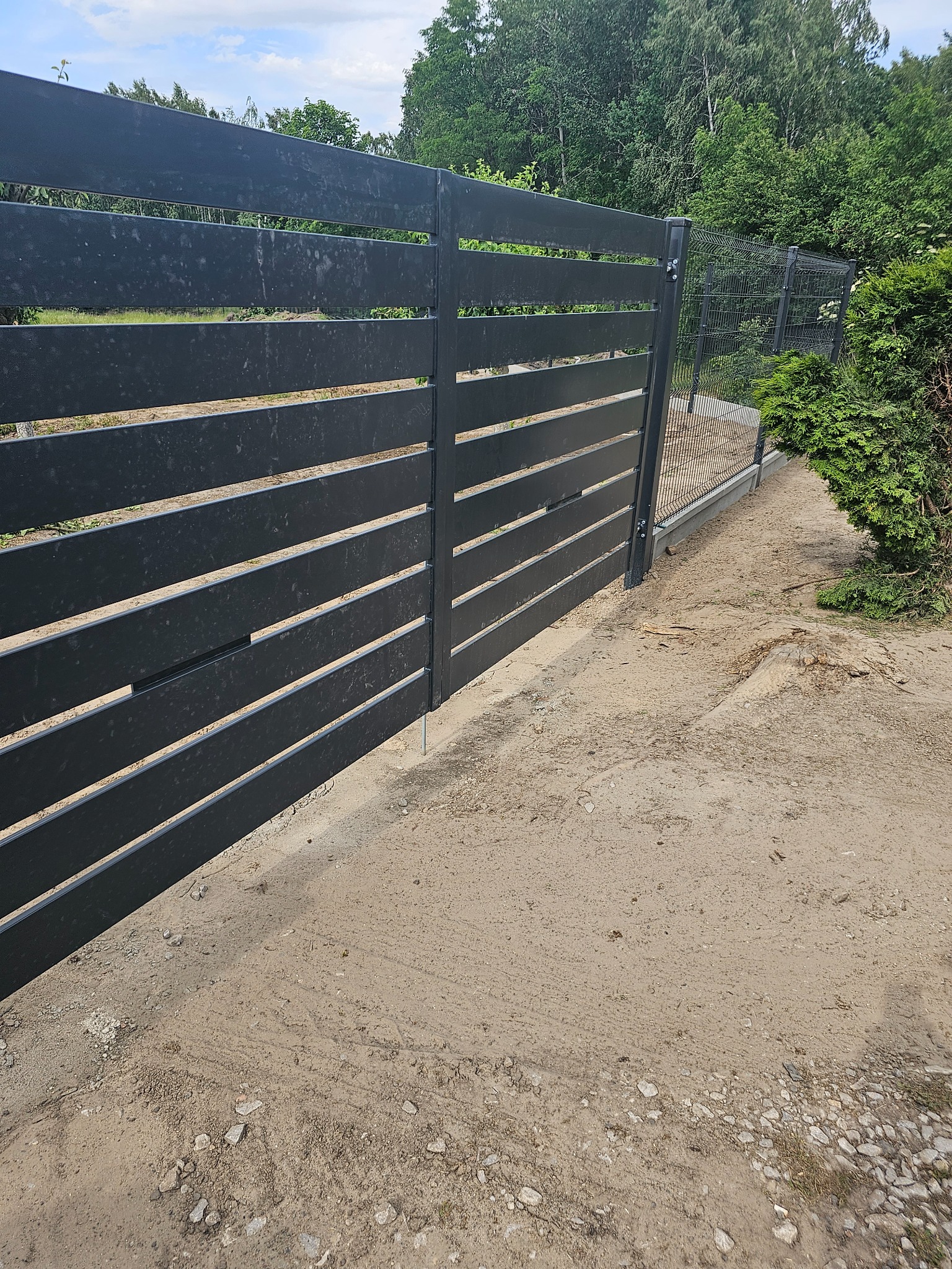 Nowoczesne, poziome ogrodzenie panelowe w kolorze grafitowym, z widoczną siatką ogrodzeniową w tle, na piaszczystym podłożu.