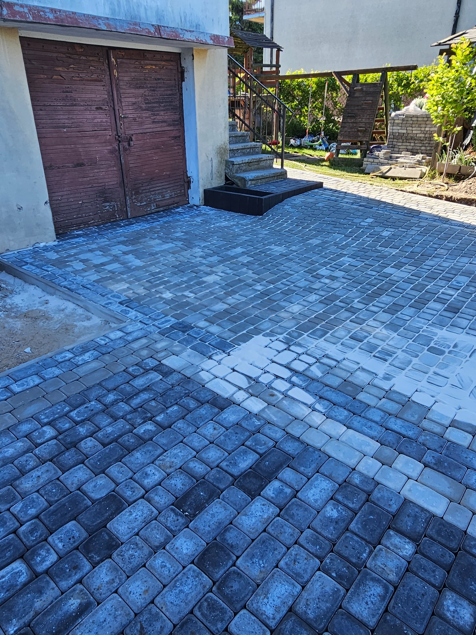 Świeżo ułożona, szara kostka brukowa przed garażem, z placem zabaw w tle. Część kostki jest jeszcze mokra, co podkreśla jej kolor i fakturę.