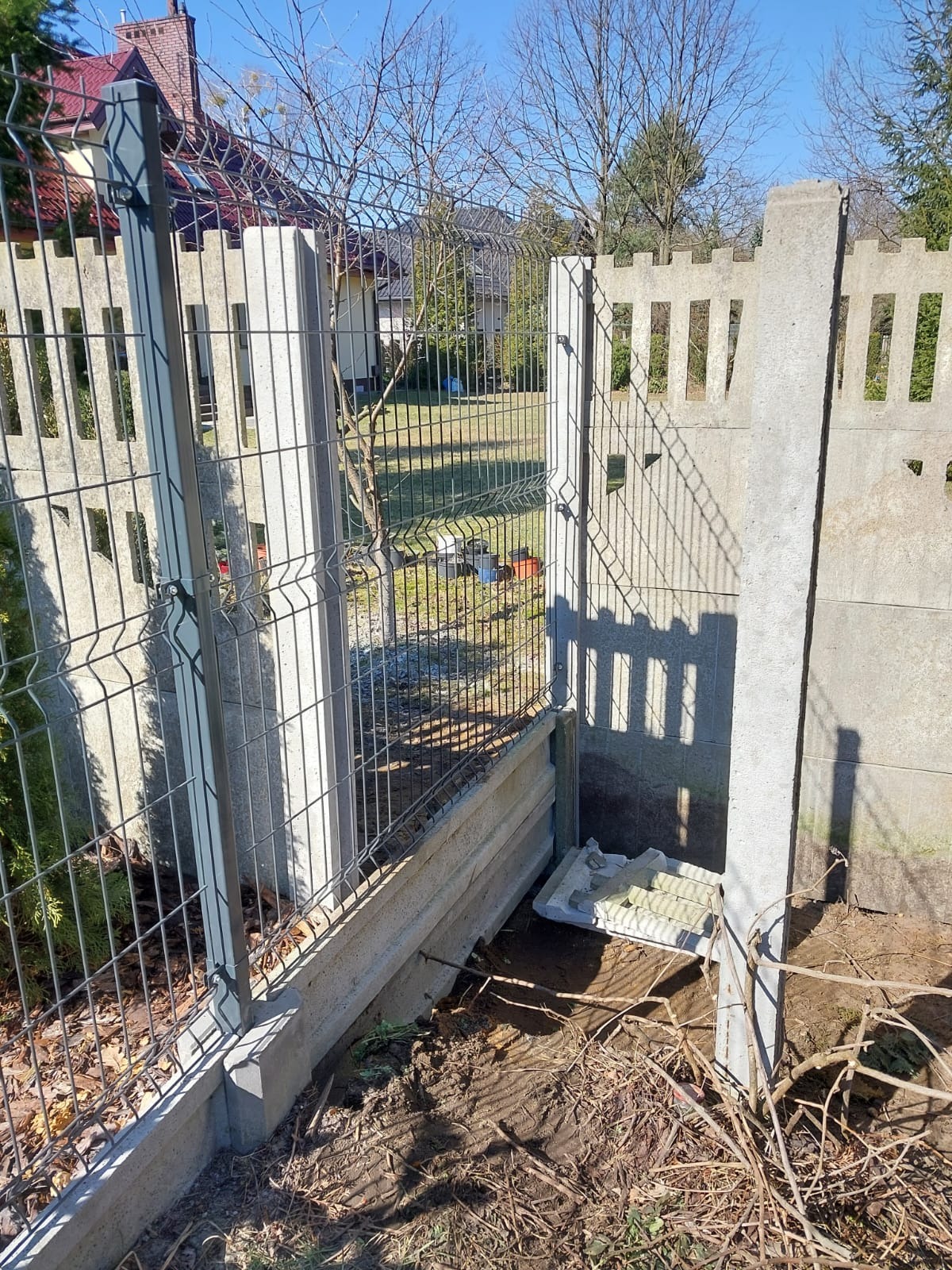 Montaż ogrodzenia panelowego z podmurówką betonową, widoczne elementy mocujące i pozostałości po pracach ziemnych, w tle dom jednorodzinny i drzewa bez liści.