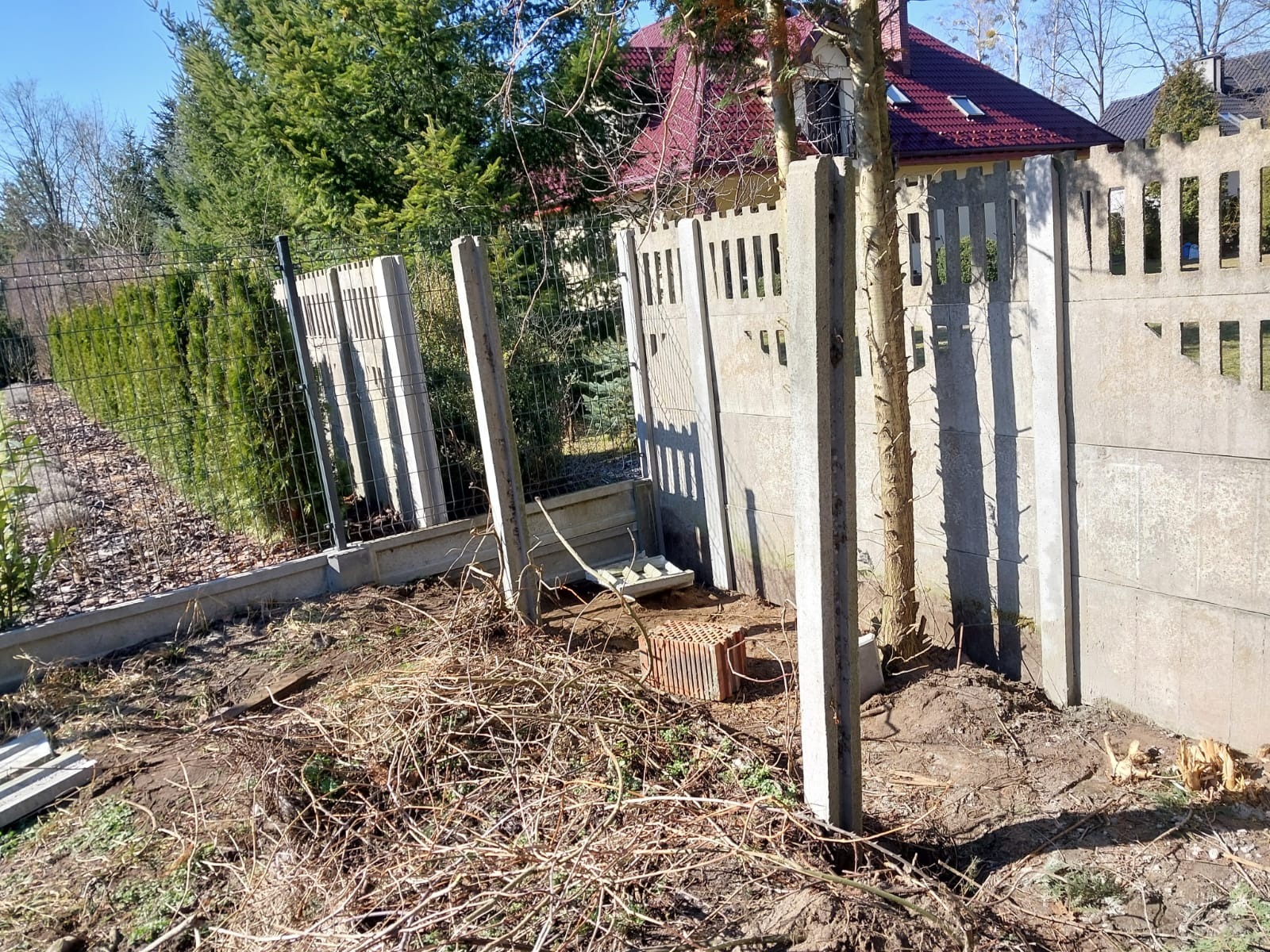 Demontaż betonowego ogrodzenia panelowego z prefabrykatów, widoczne słupki i przęsła, w tle siatka ogrodzeniowa i zieleń.