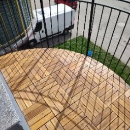 Usługi remontowo-wykończeniowe - Ułożona podłoga z drewnianych desek na balkonie, widok z góry, czarne metalowe barierki, na zewnątrz widoczny samochód dostawczy.