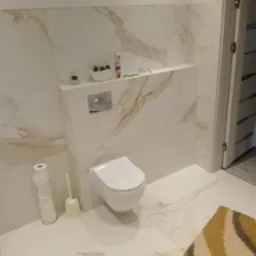 Nowoczesna łazienka z wiszącą toaletą, wykończona białymi płytkami imitującymi marmur z delikatnymi złotymi żyłkami, mała półka nad toaletą z dekoracjami, widoczny fragment dywanu w kolorach ziemi.