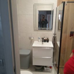 Usługi remontowo-wykończeniowe - Wykończona łazienka z beżowymi płytkami, białą szafką pod umywalką, toaletą z szarą obudową i kabiną prysznicową z drewnopodobnymi panelami, widoczny refleks fotografa w lustrze.