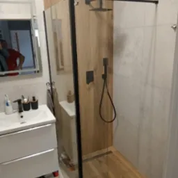 Nowoczesna łazienka z kabiną prysznicową typu walk-in, wykończoną płytkami imitującymi drewno, czarną armaturą i umywalką z szafką w kolorze białym; odbicie osoby w lustrze.