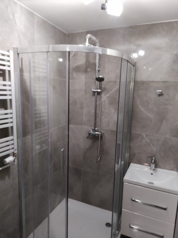 Kabina prysznicowa narożna z chromowanymi profilami i szklanymi drzwiami, umywalka z szafką w kolorze białym, grzejnik drabinkowy, szare płytki imitujące marmur na ścianach.