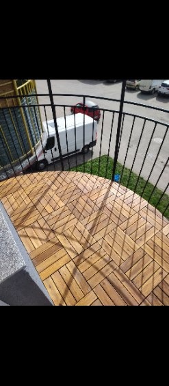 Ułożona podłoga z drewnianych desek na balkonie, widok z góry, czarne metalowe barierki, na zewnątrz widoczny samochód dostawczy.