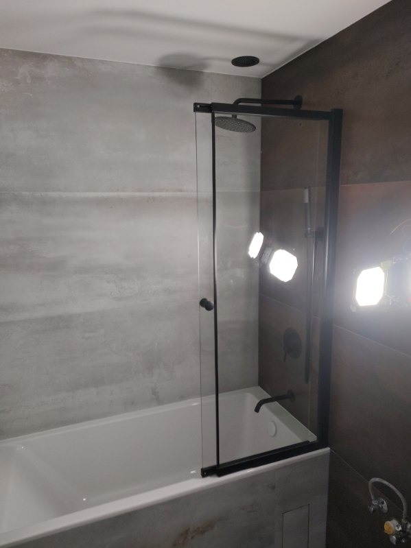 Zamontowana szklana kabina nawannowa z czarnymi profilami w łazience wykończonej płytkami w odcieniach szarości i brązu.