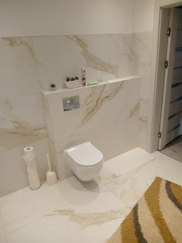 Nowoczesna łazienka z wiszącą toaletą, wykończona białymi płytkami imitującymi marmur z delikatnymi złotymi żyłkami, mała półka nad toaletą z dekoracjami, widoczny fragment dywanu w kolorach ziemi.